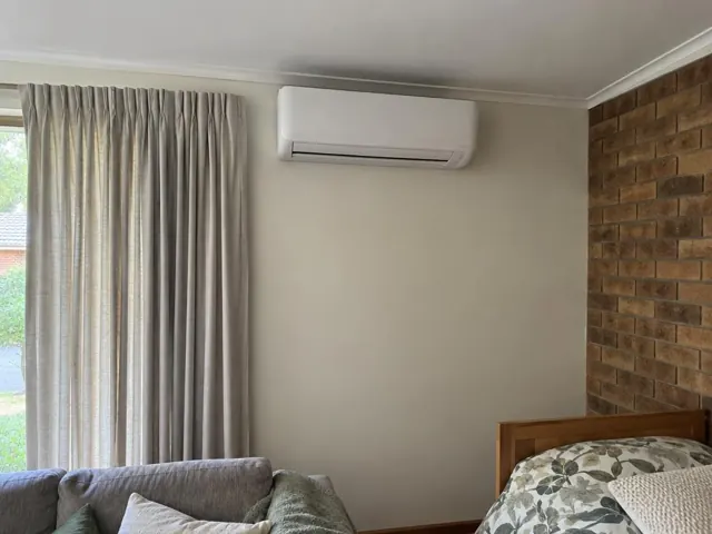 Wall-mounted split system above a sofa in a living room with curtains and brick feature wall in Happy Valley
